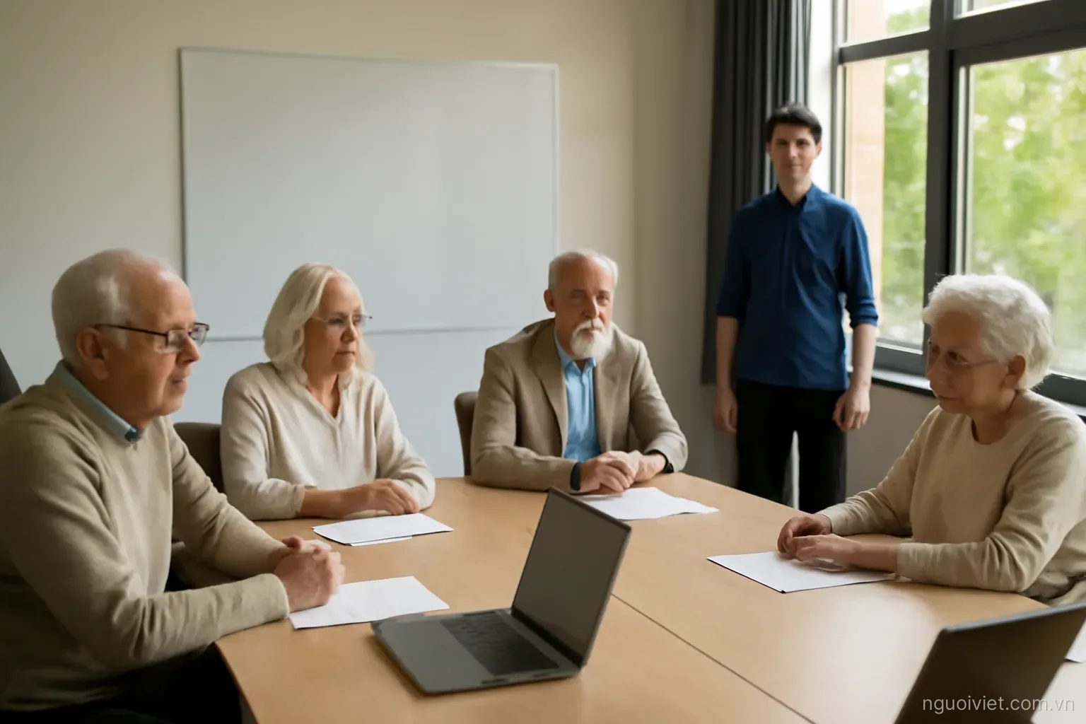 a meeting room where older people sit at a long table and a young person stands near the door